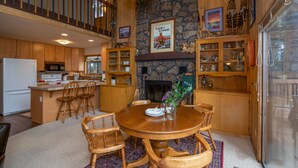 A spacious kitchen with wooden cabinets, a stone fireplace, and a dining area with a round table and chairs. - Mink #8 Close to SHARC and 8 SHARC Passes! Close to Village! (Sunriver)