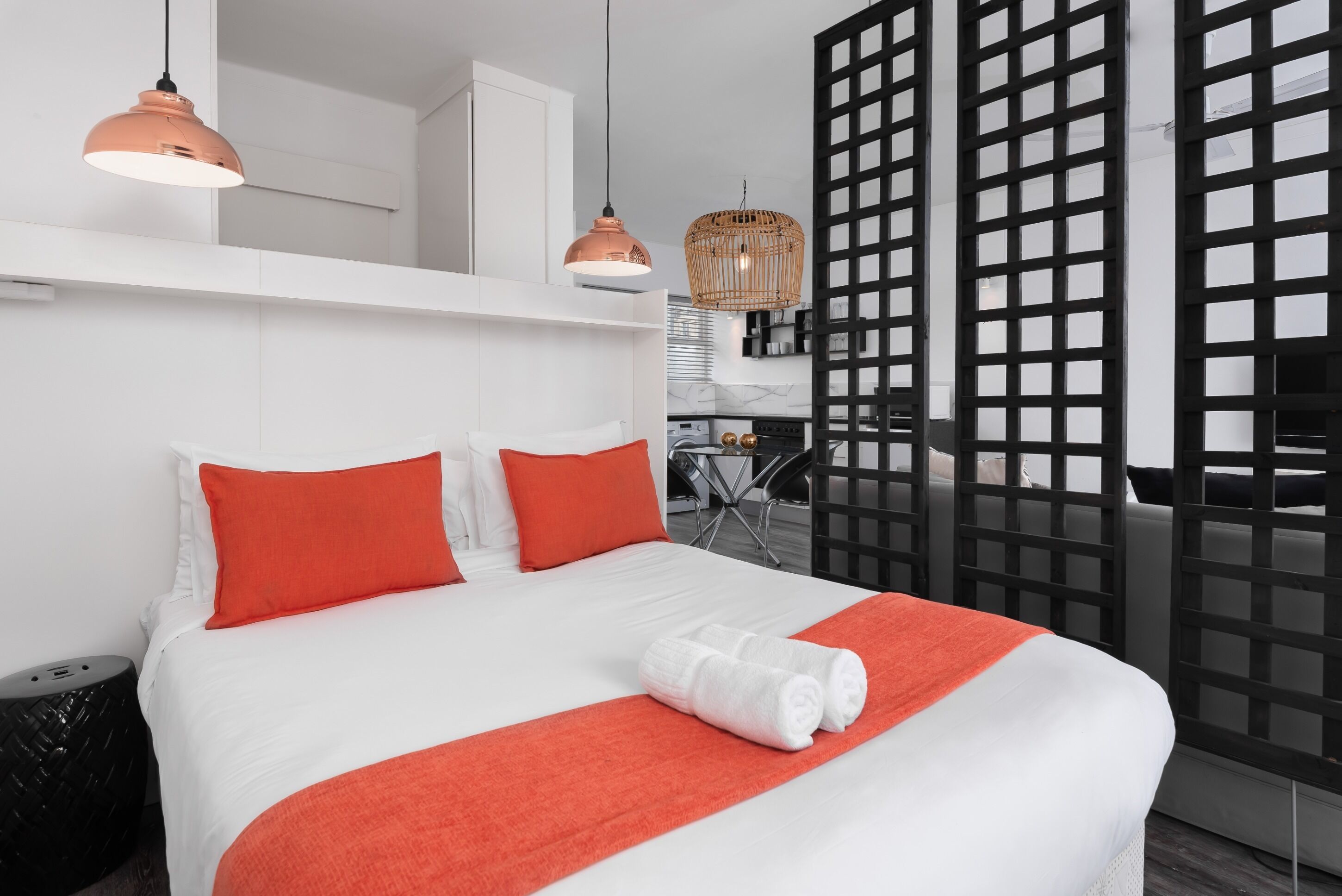 Una habitación de hotel moderna con una cama grande, almohadas rojas, una manta blanca y una cama bien hecha con toallas. Hay luces colgantes y una cesta de mimbre que cuelga del techo.