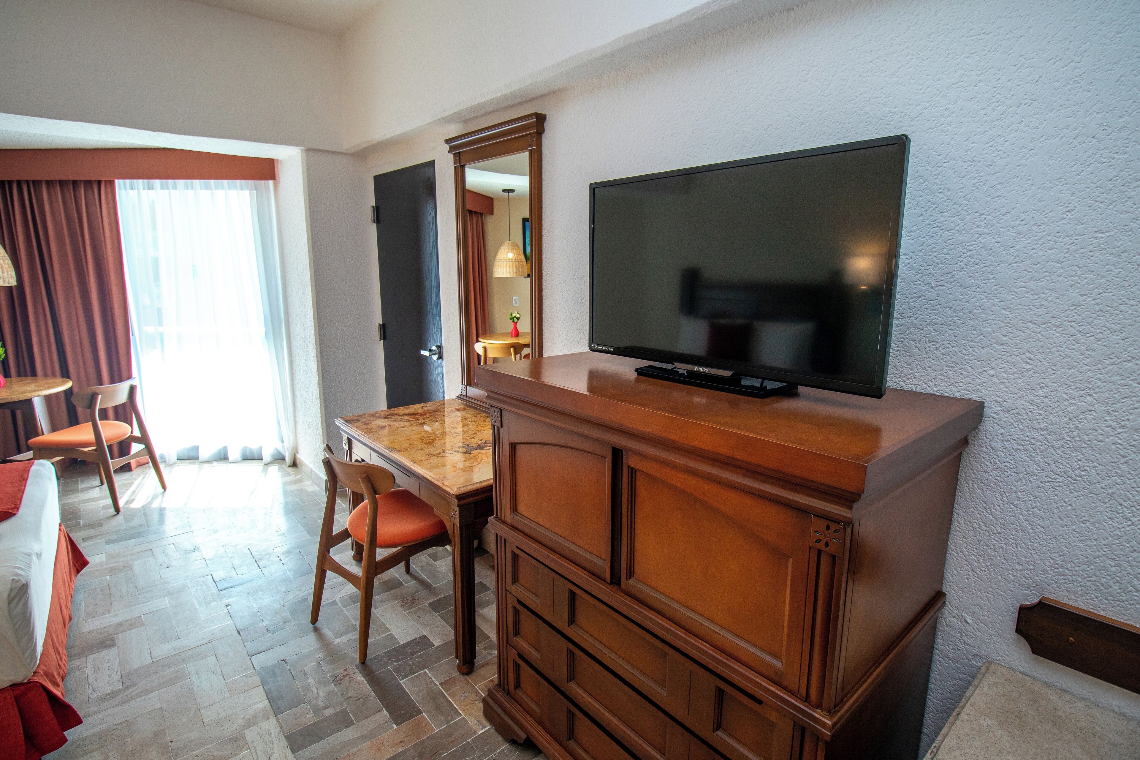 Une chambre d’hôtel avec un bureau en bois, une télévision, un miroir et un coin repas comprenant une table et des chaises.