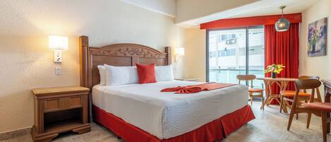 Quarto de hotel com uma cama grande, duas mesas de cabeceira, uma escrivaninha com cadeiras e uma janela com cortinas vermelhas.