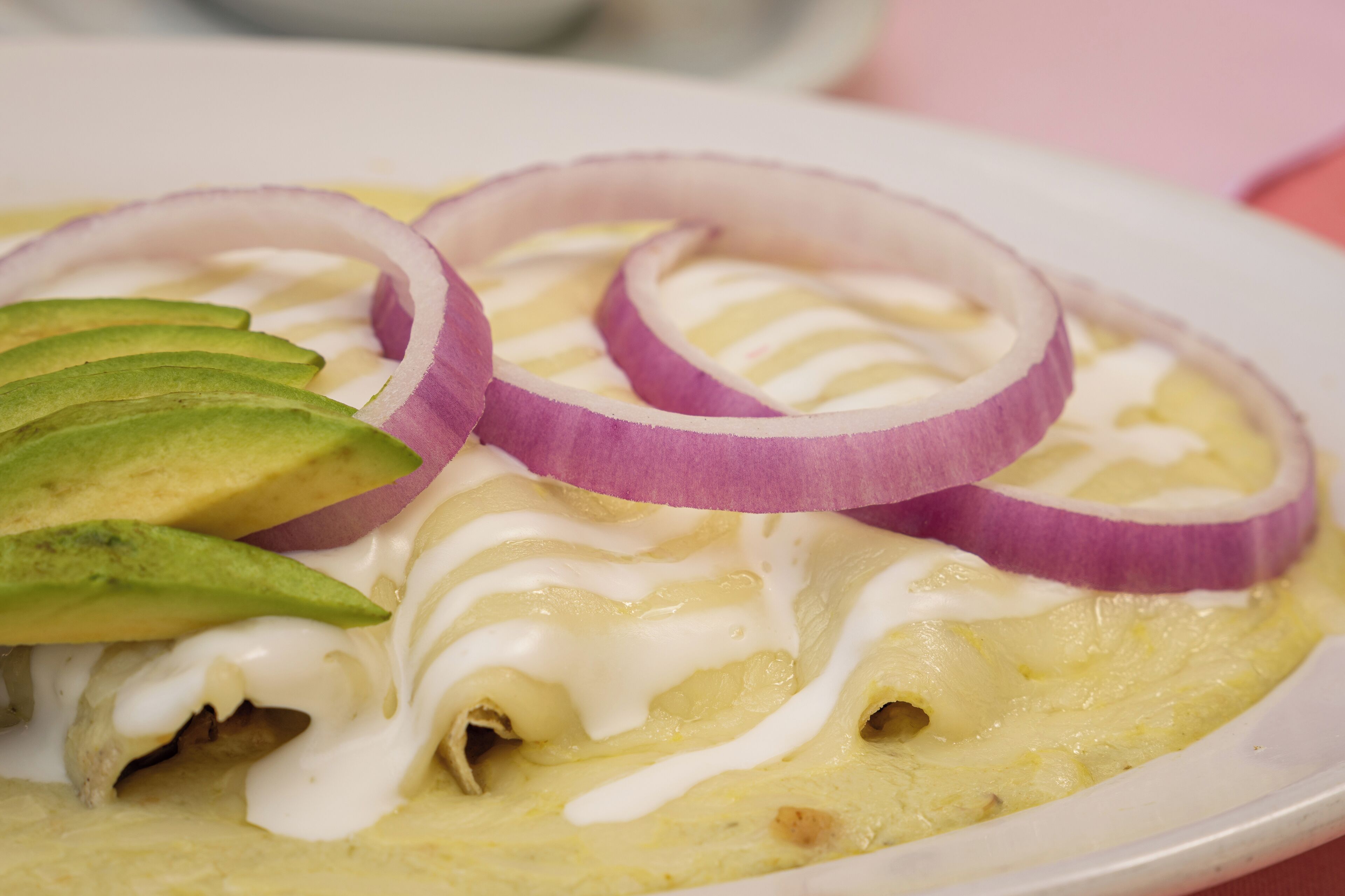 A plate of enchiladas topped with avocado slices and red onion rings, covered in a white sauce.