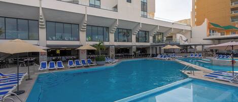 A hotel pool area with multiple lounge chairs and umbrellas, surrounded by modern buildings.