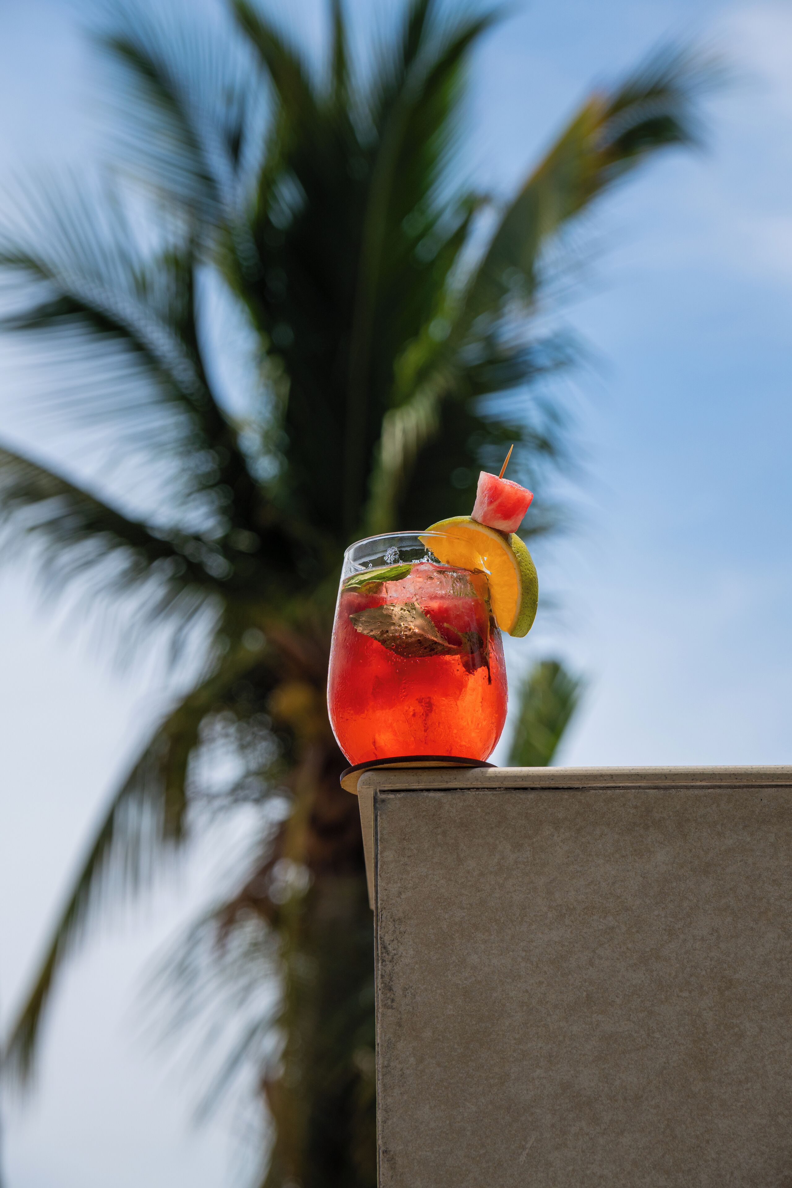 A glass of red beverage with a slice of lime and a strawberry on a table with a palm tree in the background.