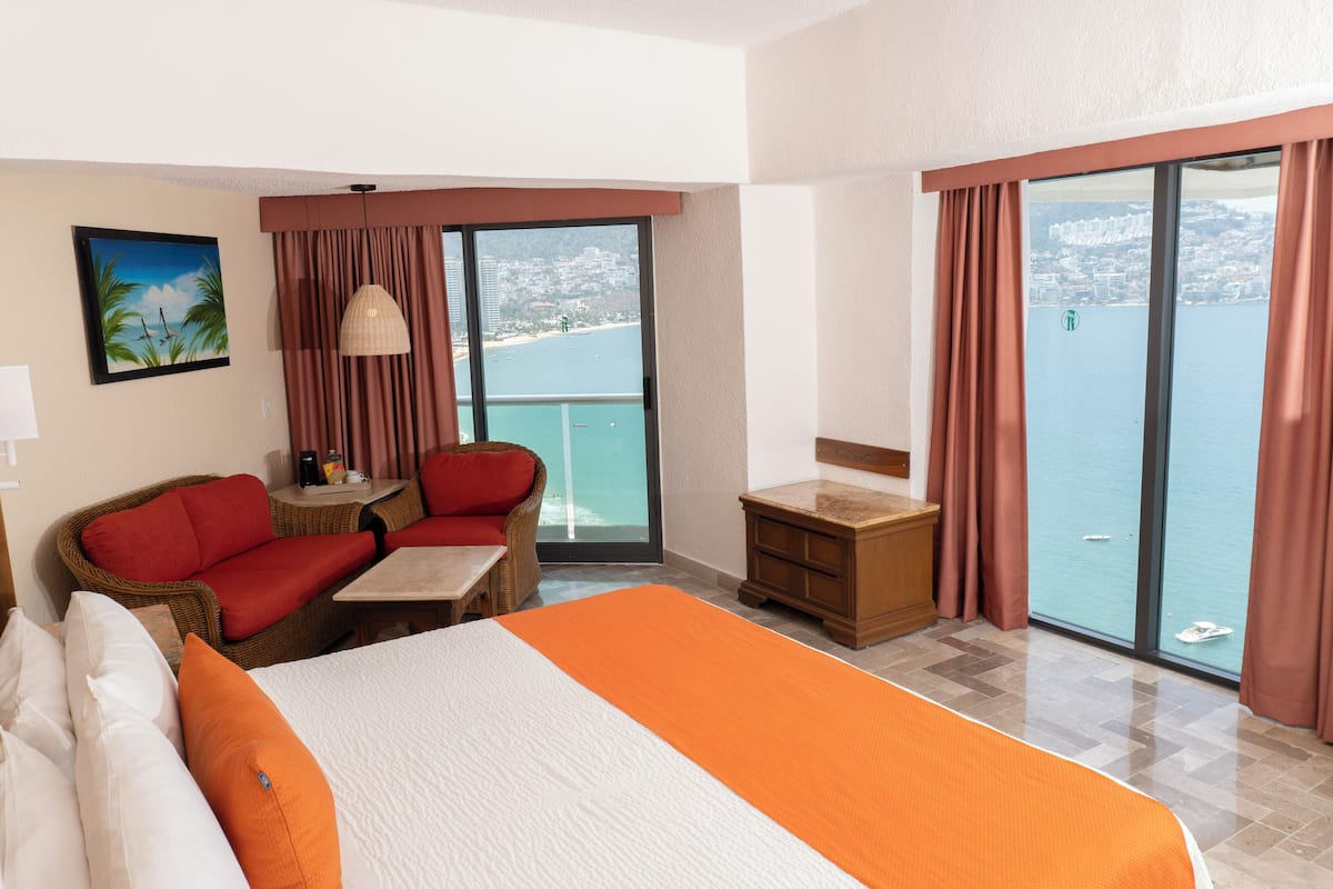 Une chambre d’hôtel comprenant un lit, un coin salon avec une table et des chaises, un petit bureau et une grande fenêtre donnant sur la mer et les bâtiments.