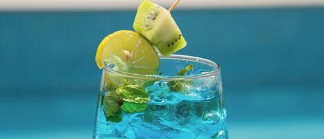 A glass of blue beverage with lime and mint garnish, placed on a table by a pool.