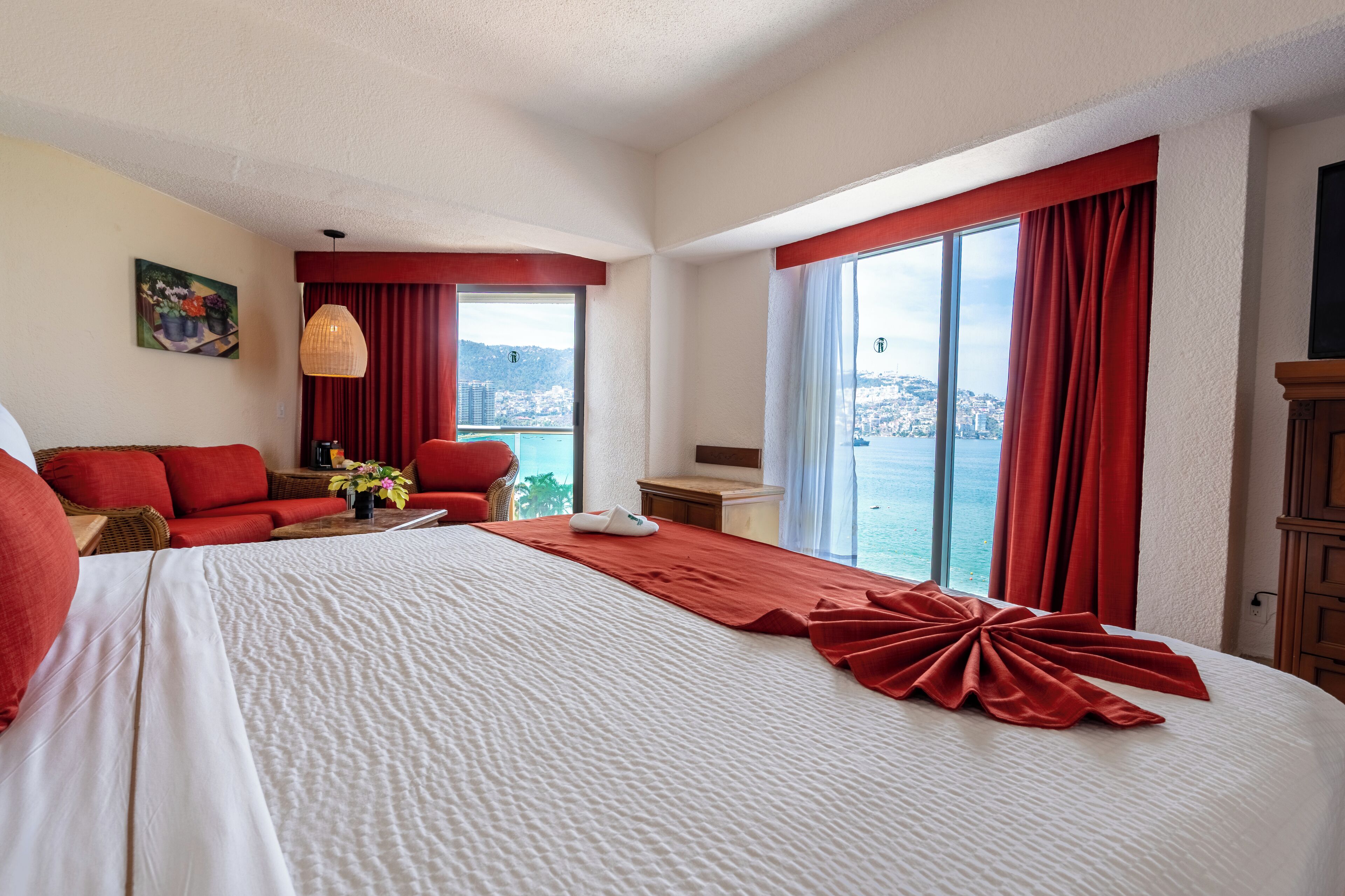 Une chambre d’hôtel avec un grand lit, des rideaux rouges, une vue sur la mer, un canapé et un tableau au mur.