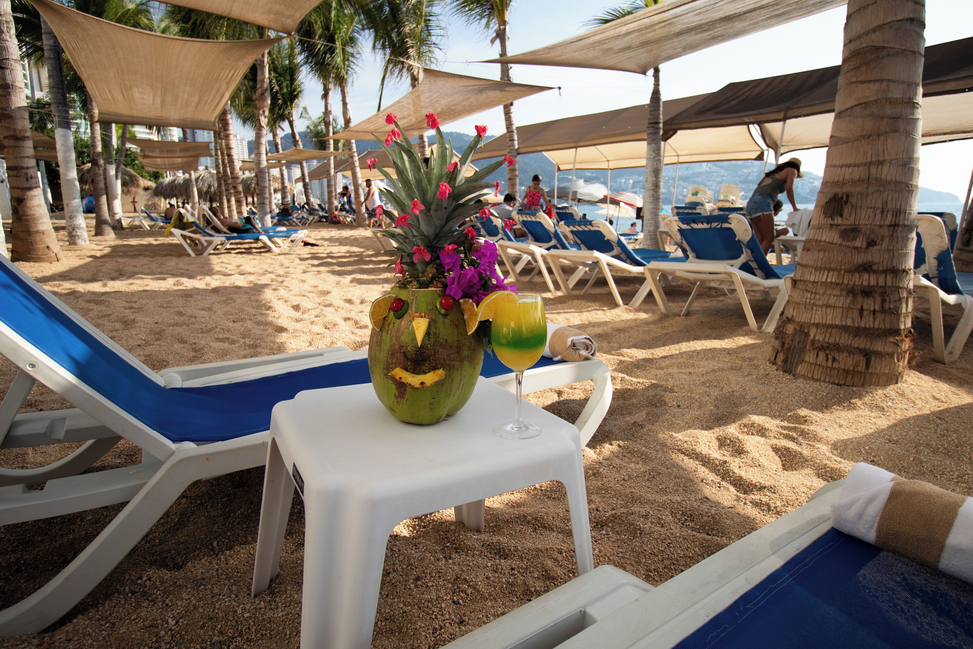 Una playa con tumbonas, una mesa con un coco y flores, y palmeras.