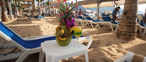 A beach with sun loungers, a table with a coconut and flowers, and palm trees.