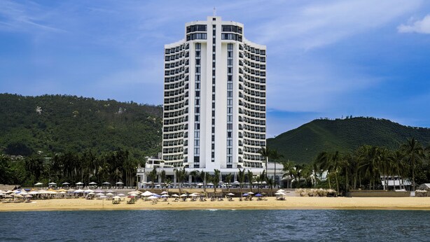 Un hotel alto con una torre central, ubicación frente al mar y sombrillas de playa.
