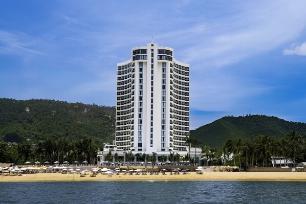 Un hotel alto con una torre central, ubicación frente al mar y sombrillas de playa.