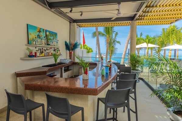 A bar area with a curved granite countertop, black bar stools, and a view of the ocean. There are potted plants and a painting on the wall.