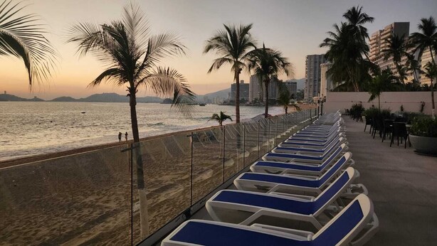 Un hotel frente a la playa con filas de tumbonas azules y blancas a lo largo de la costa, palmeras y un horizonte urbano al fondo.