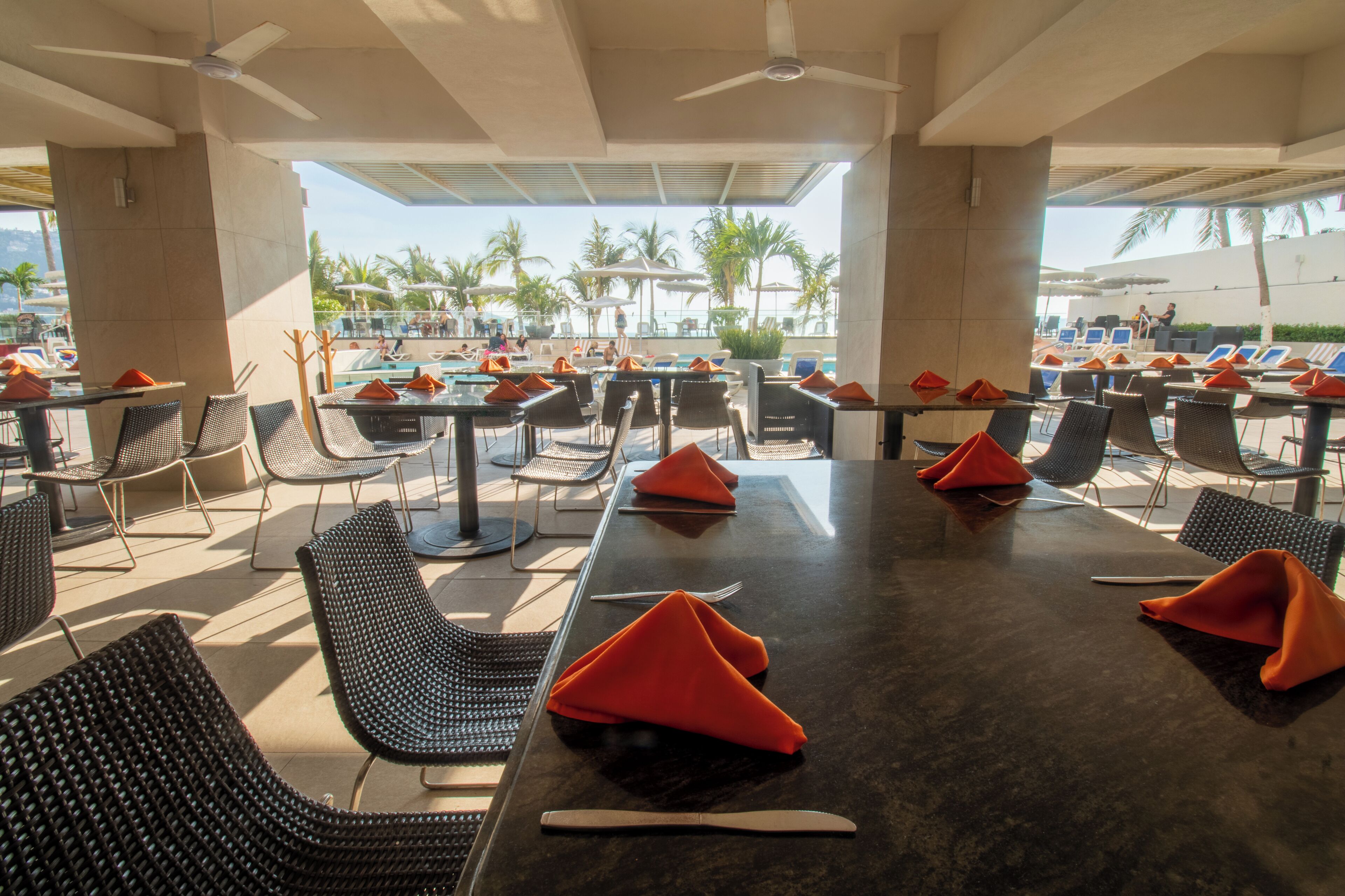 A restaurant with outdoor seating, tables, chairs, and umbrellas, overlooking a pool area.
