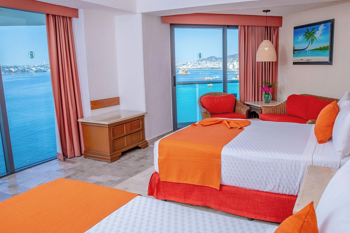 Une chambre d’hôtel dotée d’une grande fenêtre donnant sur la mer et d’un balcon. On y trouve un lit avec une literie orange et blanche, une table de chevet en bois et une chaise en osier avec des coussins rouges.