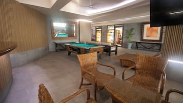 A billiards table, wicker chairs, a television, and a potted plant in a room with a tiled floor and a ceiling fan.