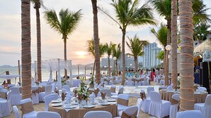 Un salón de bodas al aire libre con mesas redondas, sillas blancas y doradas, y palmeras.