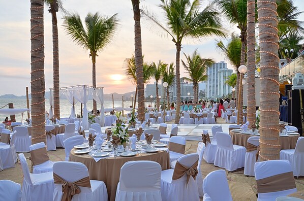 An outdoor wedding venue with round tables, white and gold chairs, and palm trees. - Copacabana Beach Hotel Acapulco (Acapulco)