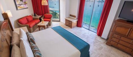 A hotel room with a large bed, a sofa, a small table, a TV, and a balcony with a view of the sea.