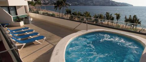 A pool with a view of the sea, lounge chairs, and a building with balconies.