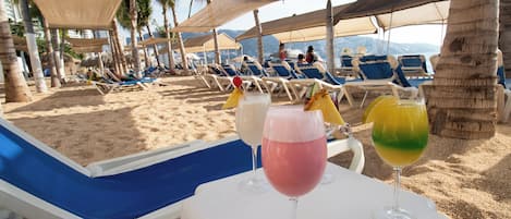 Un área de descanso en la playa con tumbonas y sombrillas, una mesa con bebidas y vistas a la playa y el océano.