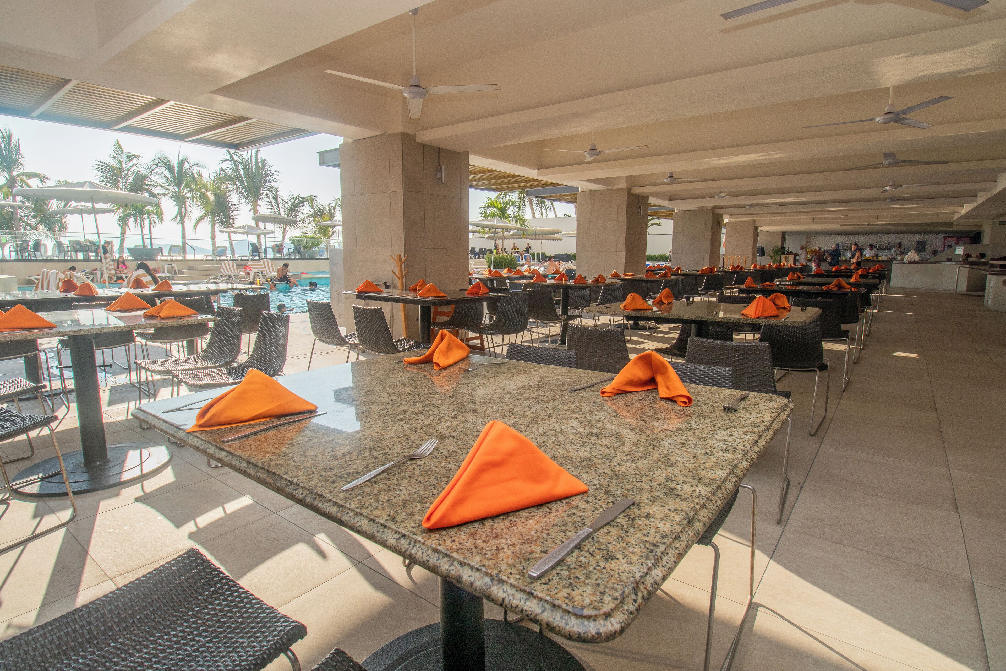Un'ampia zona pranzo all'aperto con tavoli e sedie, una piscina e palme sullo sfondo.