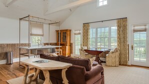A spacious living area with a wooden table, brown sofa, and a dining table with chairs. There is a kitchen area with cabinets and a window. - Ramsay Estate Carriage House – Luxury Retreat with Mountain Views (Greenwood)