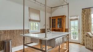 A kitchen with a central island, wooden flooring, a trash can, a window with blinds, a wooden cabinet, and a door with a curtain. - Ramsay Estate Carriage House – Luxury Retreat with Mountain Views (Greenwood)