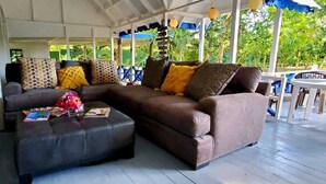 A covered outdoor seating area with a brown sofa and ottoman, surrounded by blue and white chairs.