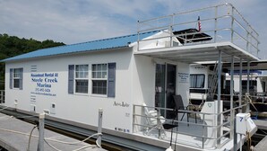 Una casa flotante con techo azul y paredes blancas, con la leyenda 'Houseboat Rental At Steele Creek Marina'.