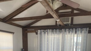 Una habitación con techo de madera, cómoda blanca, sofá color turquesa, almohada a rayas, mesa de madera y una ventana con persianas.