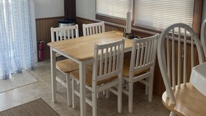 Un comedor con una mesa de madera y sillas blancas, un televisor montado en la pared y una ventana con persianas.