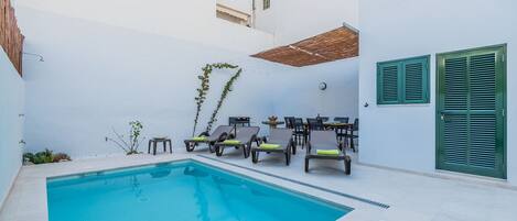 A swimming pool with lounge chairs and a table, surrounded by white walls and a green shuttered door.