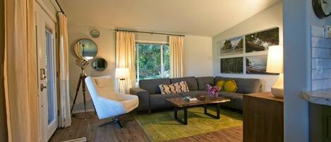Una sala de estar con sofá, sillón, mesa de centro y lámpara. Se observa una ventana con cortinas, una puerta y un reloj de pared.
