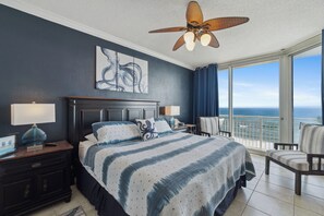 Une chambre avec un grand lit, un ventilateur de plafond, une table de chevet, une chaise et une vue sur l’océan.