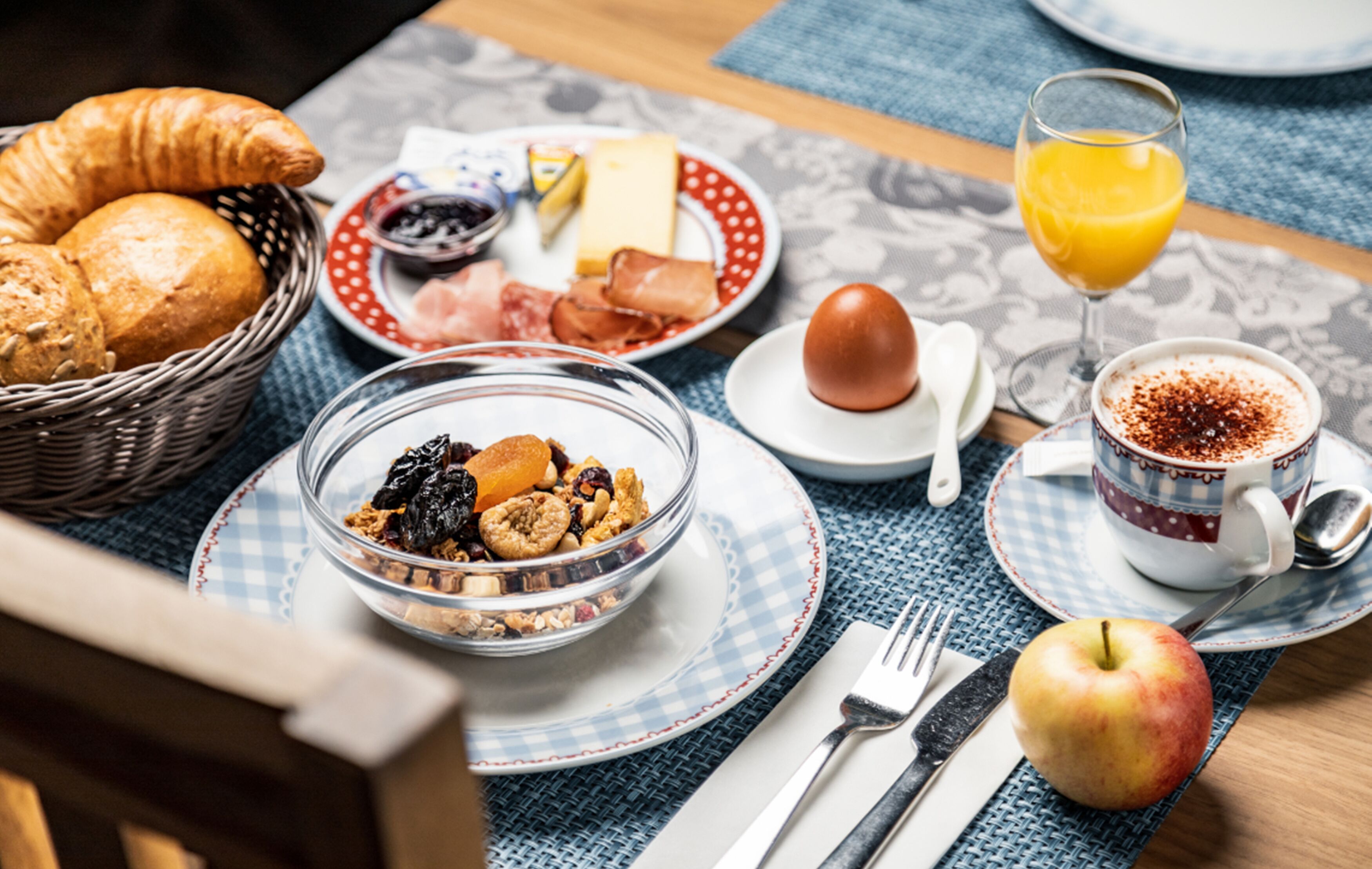 Desayuno con cruasanes, un tazón de cereal, un vaso de jugo de naranja, una taza de café y una manzana sobre una mesa.