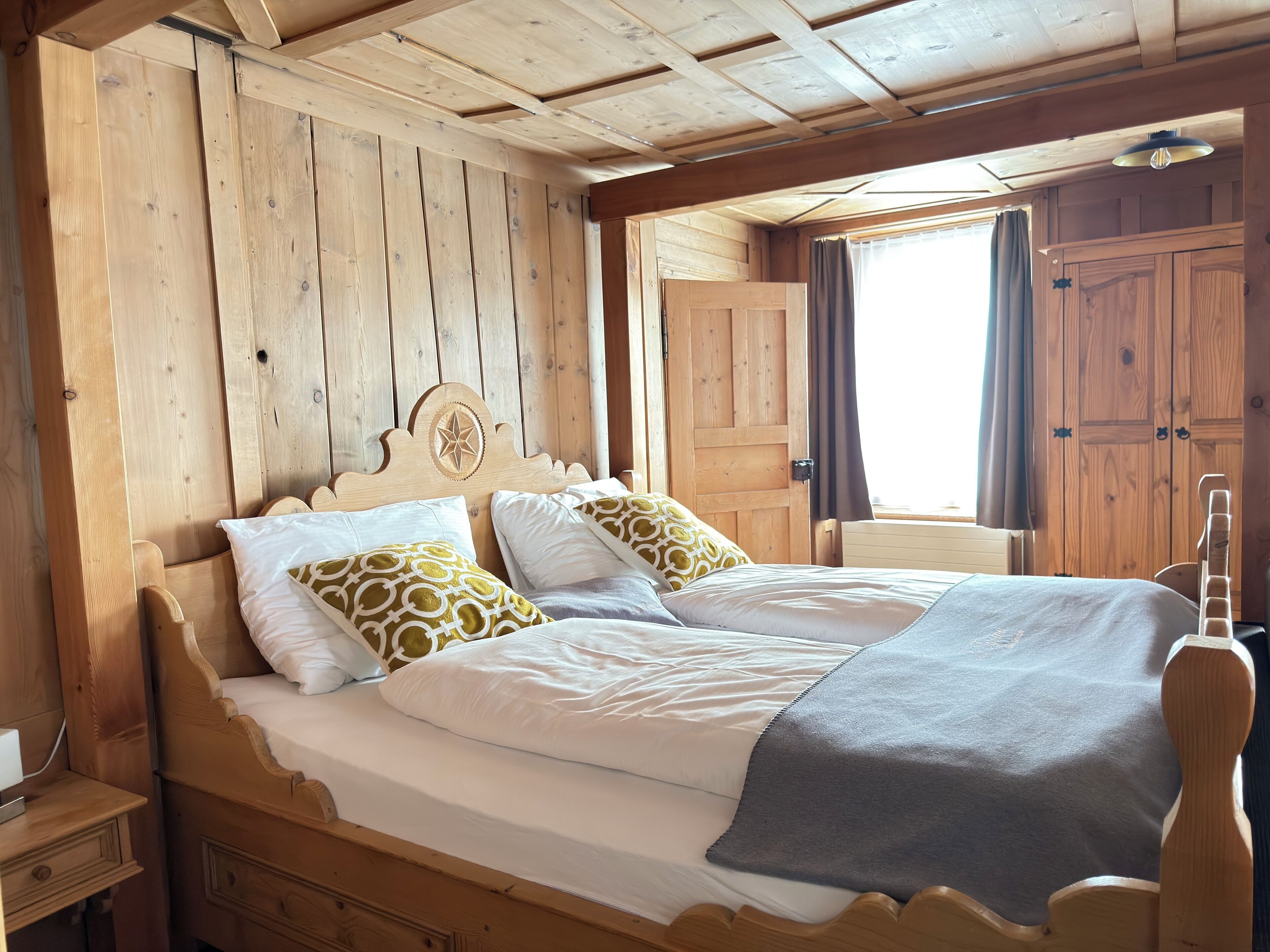 Ein hölzernes Bett mit zwei Kissen, ein Nachttisch und ein Fenster mit Vorhängen.