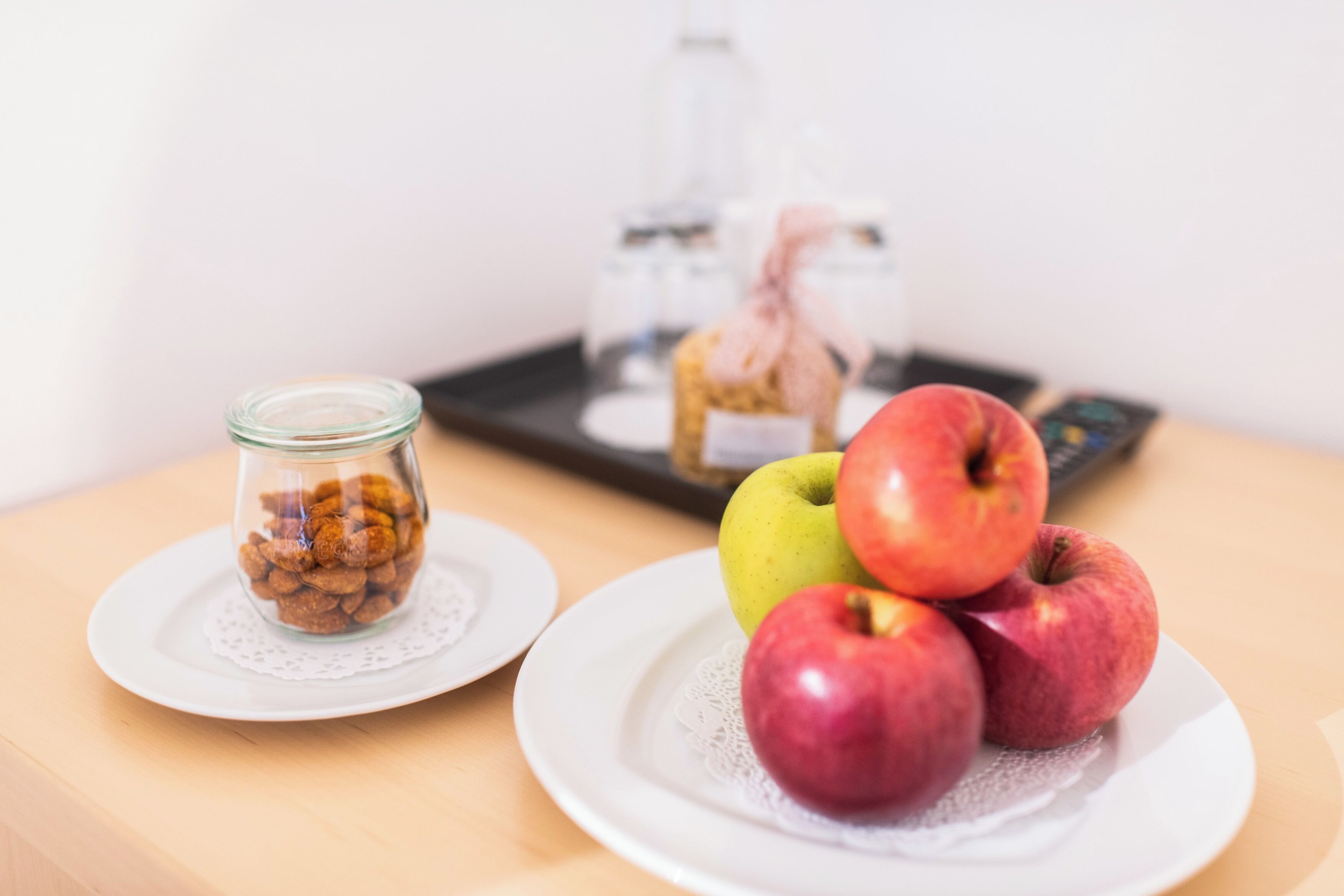 Una mesa con un tarro de cristal con nueces, un plato con tres manzanas y una pequeña botella con un lazo rosa.
