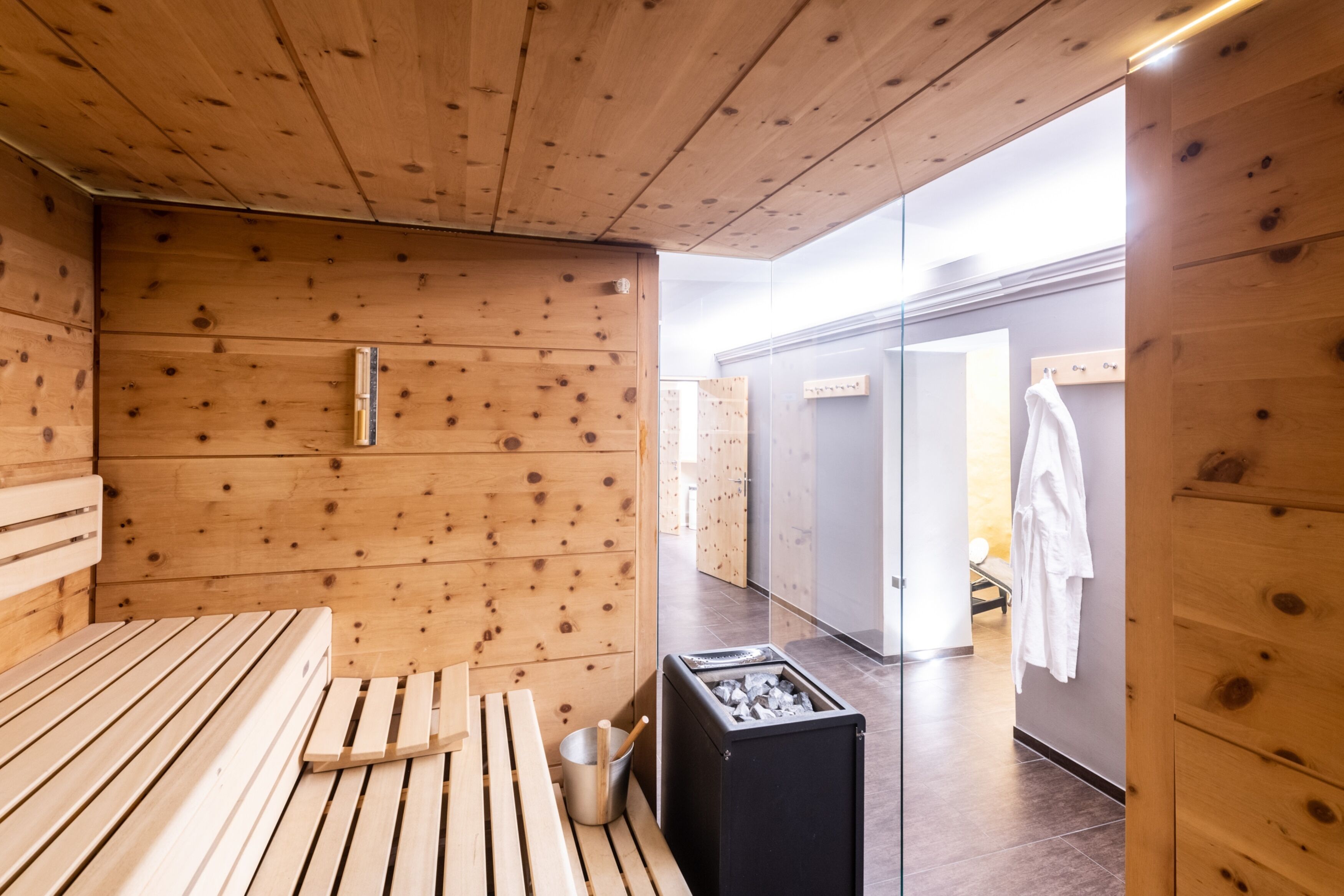 Una sala de sauna con paredes y bancos de madera, un calentador de sauna y una puerta de vidrio que da a un vestuario con un perchero.