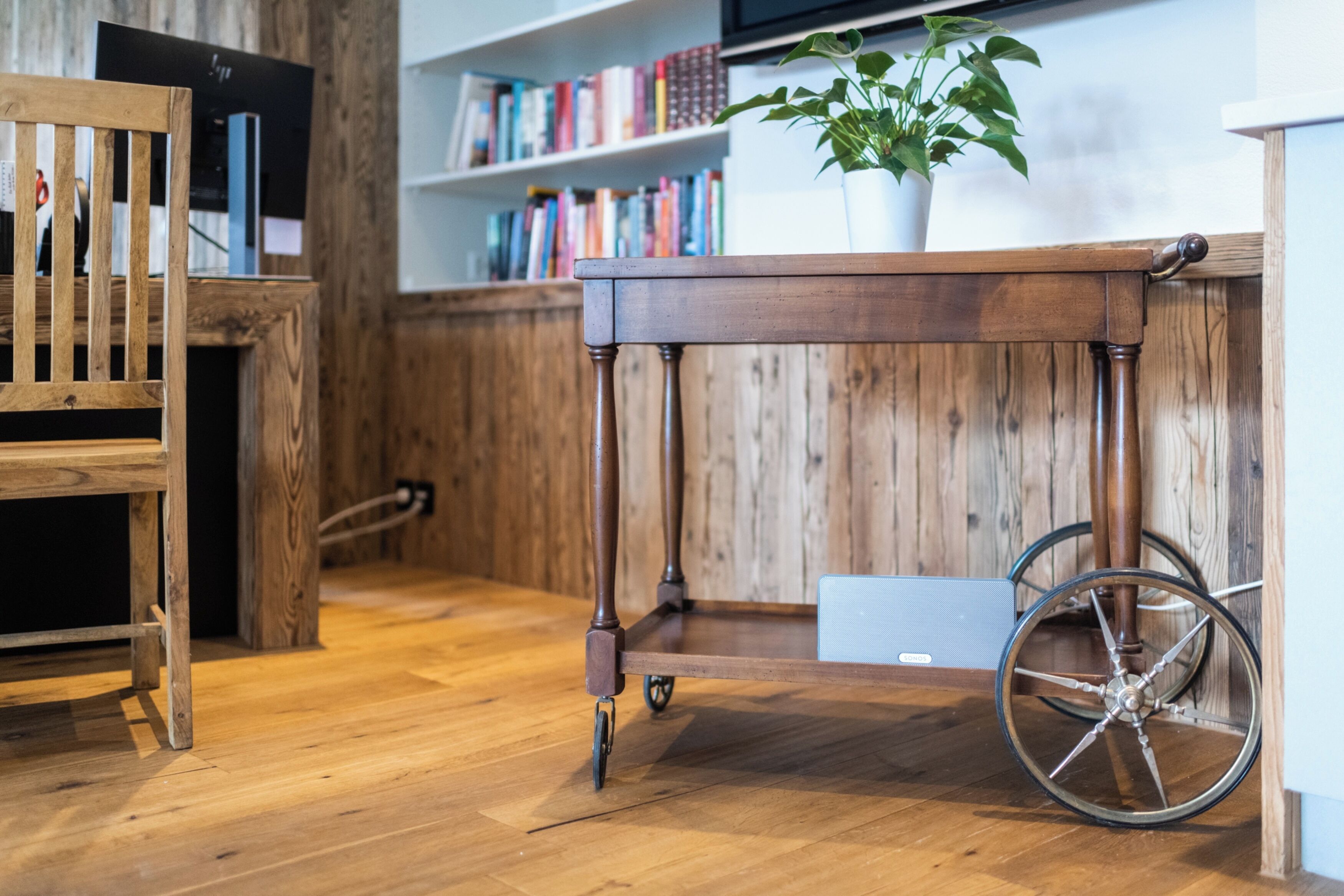 A wooden table with wheels, a wooden chair, and a bookshelf with books in a room with wooden flooring.