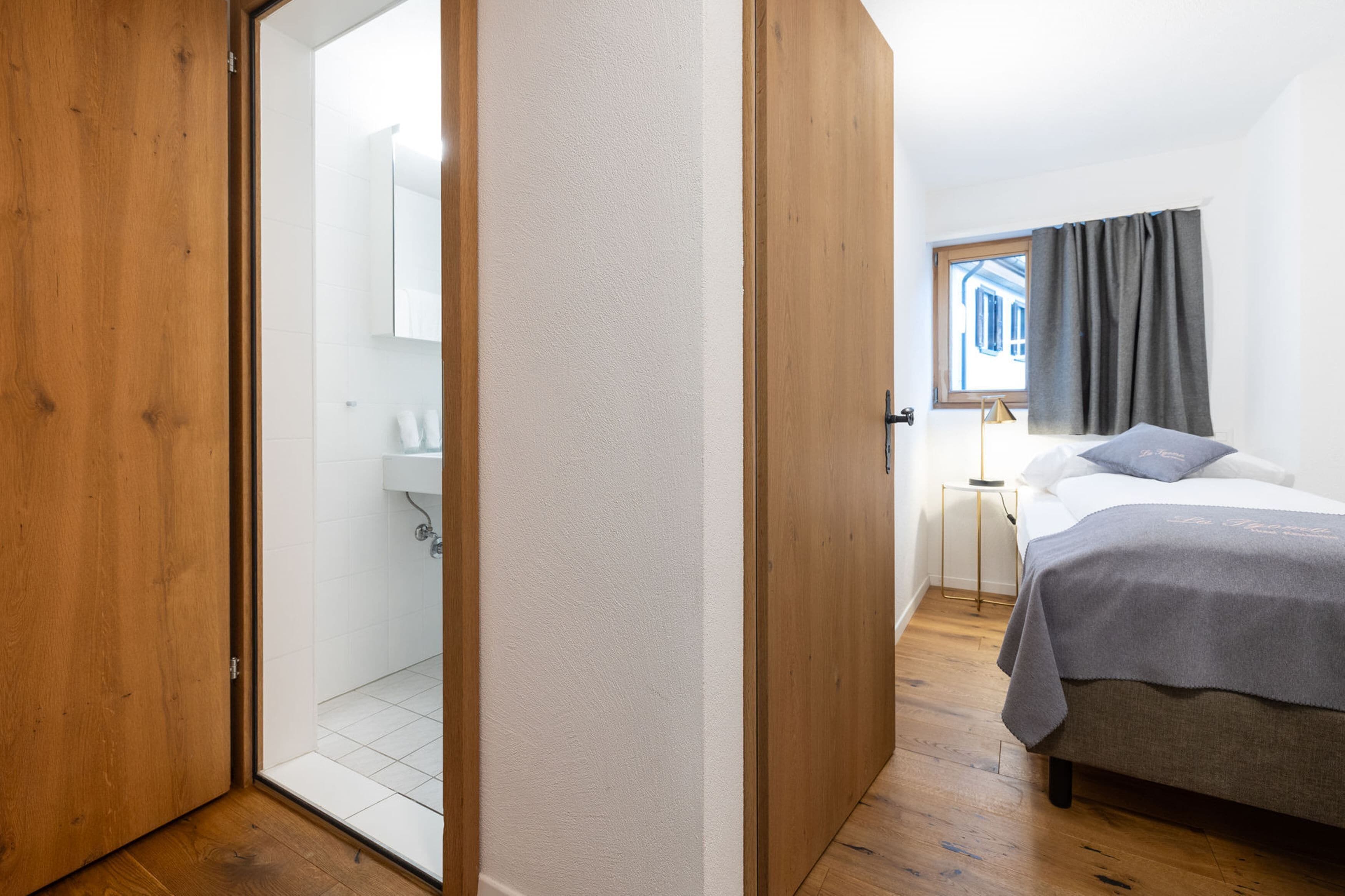 Ein Schlafzimmer mit Bett, Nachttisch, einem Fenster mit Vorhängen und Blick auf ein Gebäude.