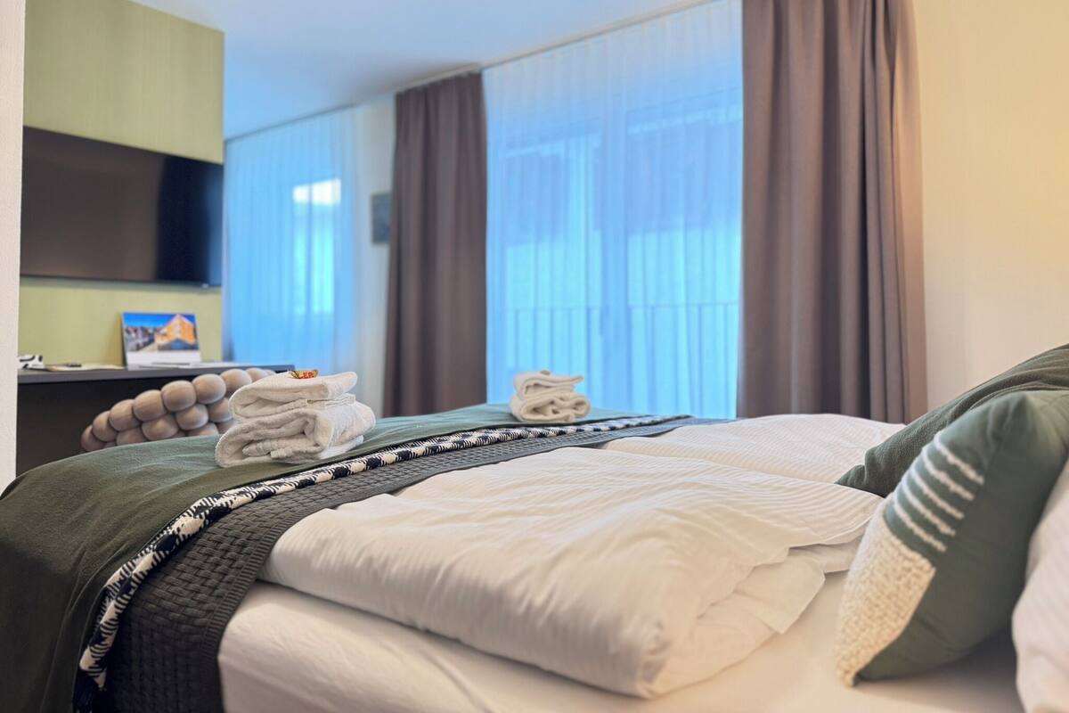 Hotellihuone, jossa on sänky, työpöytä tuolilla, televisio ja ikkuna, jossa on verhot.