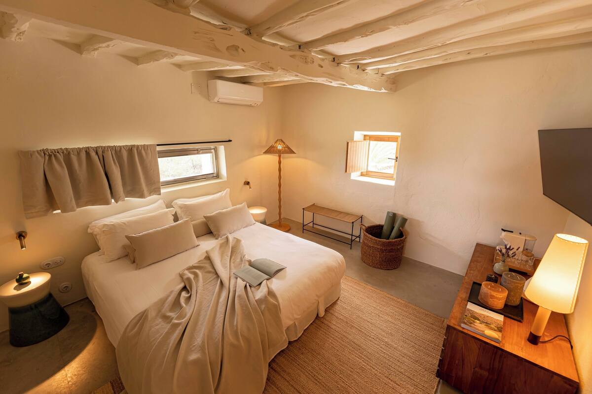 Une chambre avec un grand lit, une table de chevet, une lampe et une petite fenêtre.