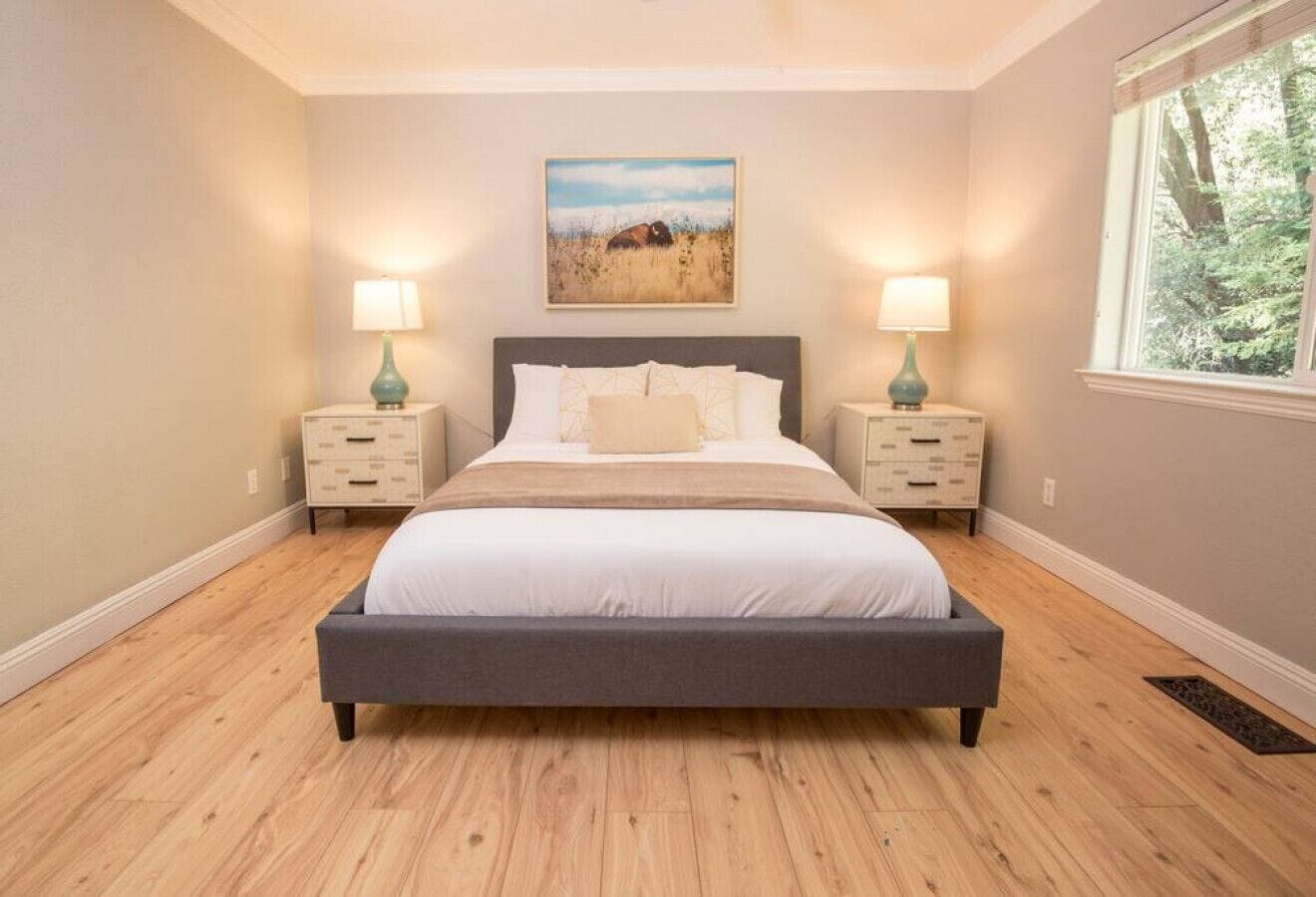A bedroom with a large bed, two bedside tables with lamps, a painting on the wall, and a window with a view of trees.