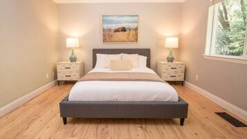 A bedroom with a large bed, two bedside tables with lamps, a painting on the wall, and a window with a view of trees.