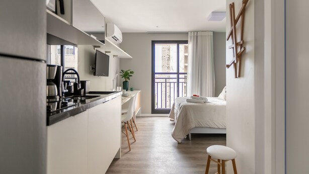 A compact hotel room with a kitchenette, a bed, a small seating area, and a balcony with a view.