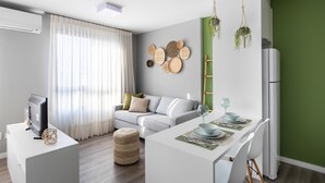 A modern living room with a sofa, a dining table set with plates and glasses, and a kitchen area with a refrigerator and hanging plants.