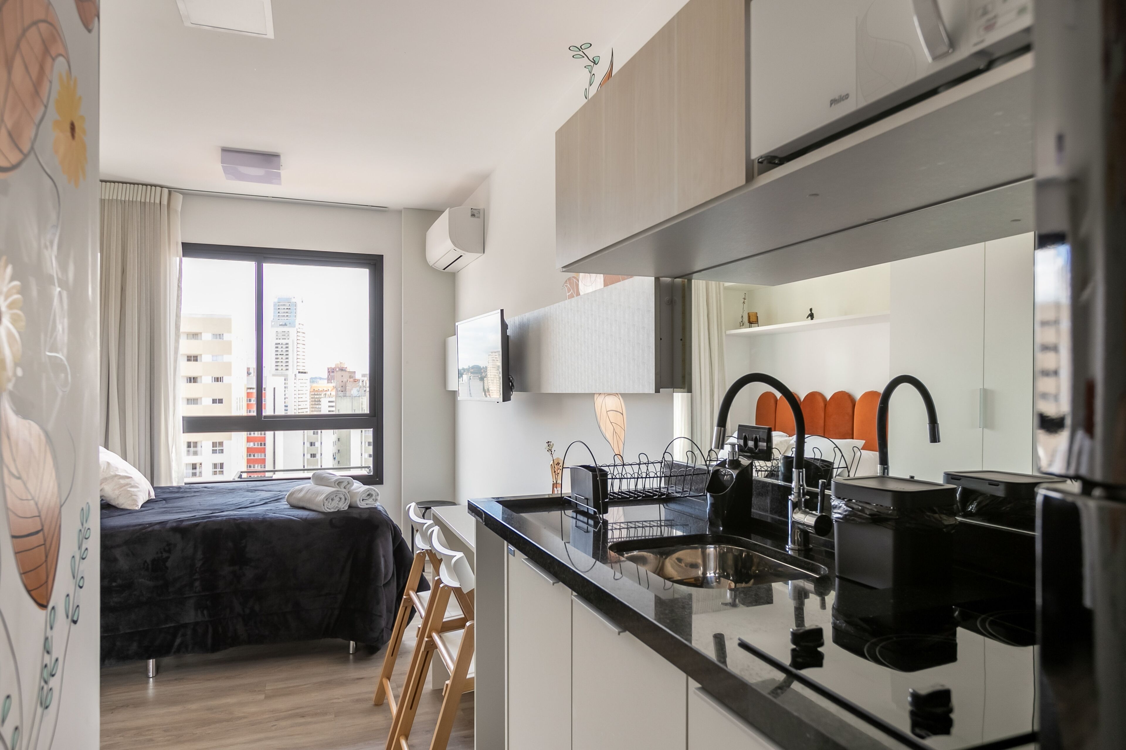 A modern hotel room with a bed, kitchenette, and a large window offering a city view.