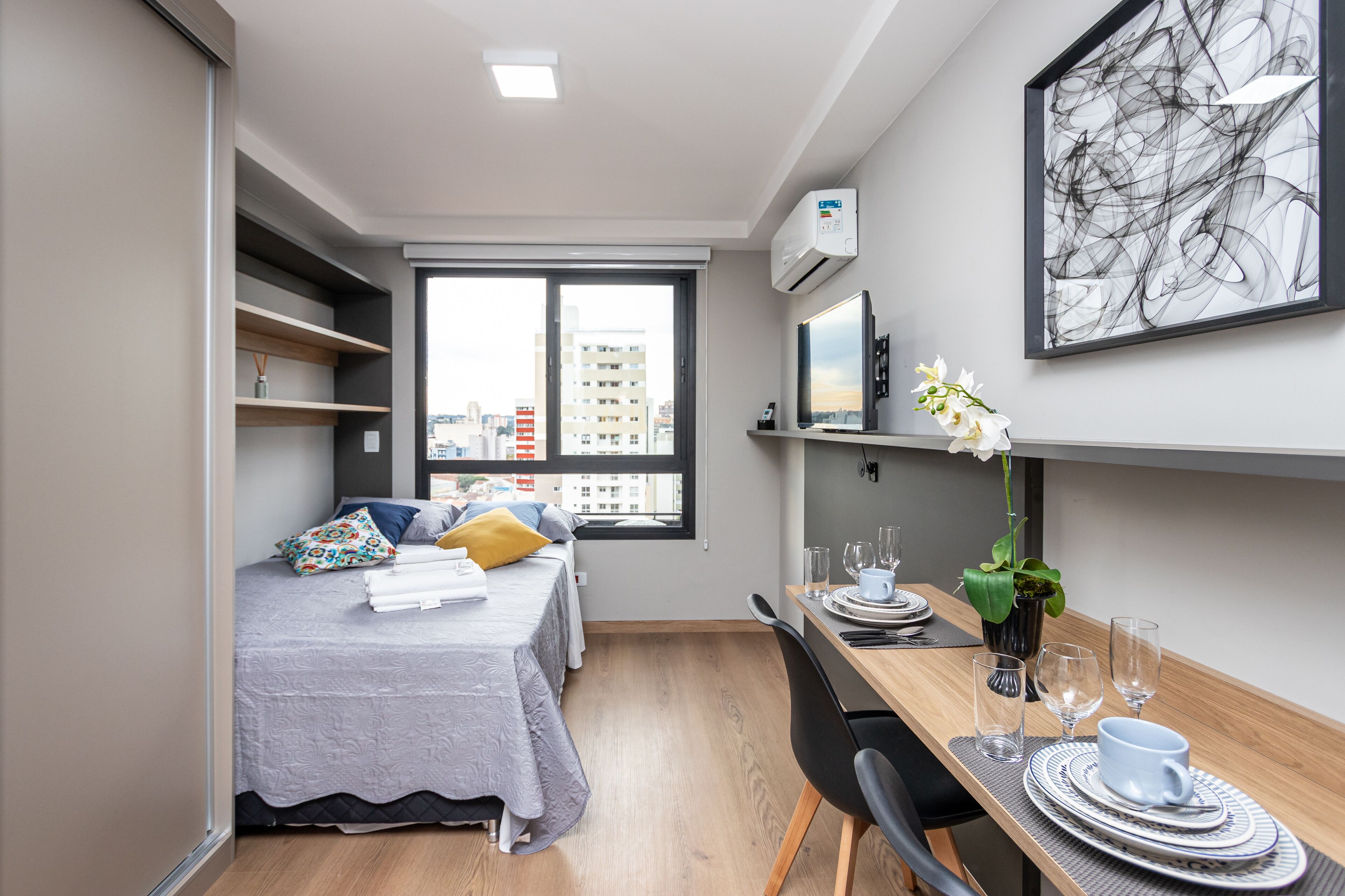 Ein kompaktes Hotelzimmer mit einem Bett, einem Esstisch für zwei Personen, einem kleinen Fenster mit Blick auf die Stadt und einer Wand-Klimaanlage.