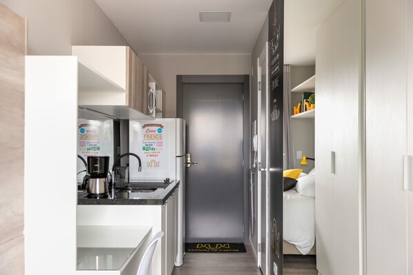 A compact kitchen with a refrigerator, microwave, and coffee maker. A door with a doormat is visible. - All You Need (Curitiba)
