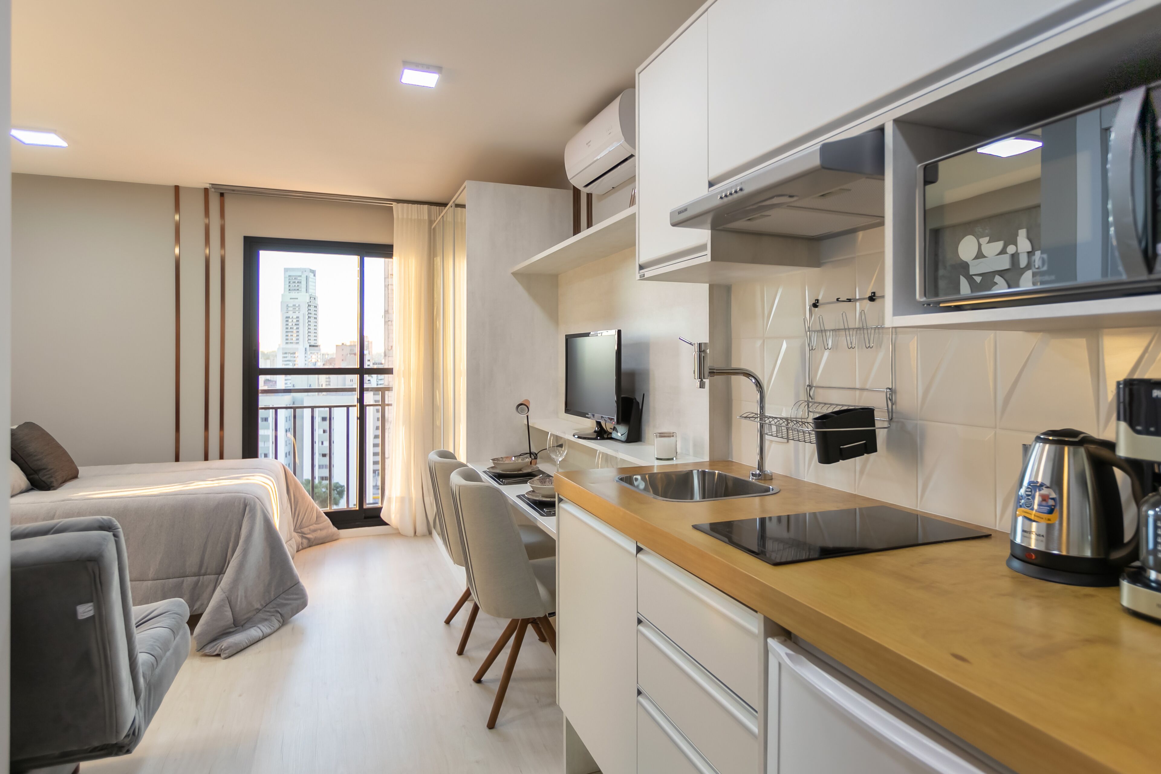 A compact hotel room with a kitchenette, a bed, a dining area, and a large window offering a city view.